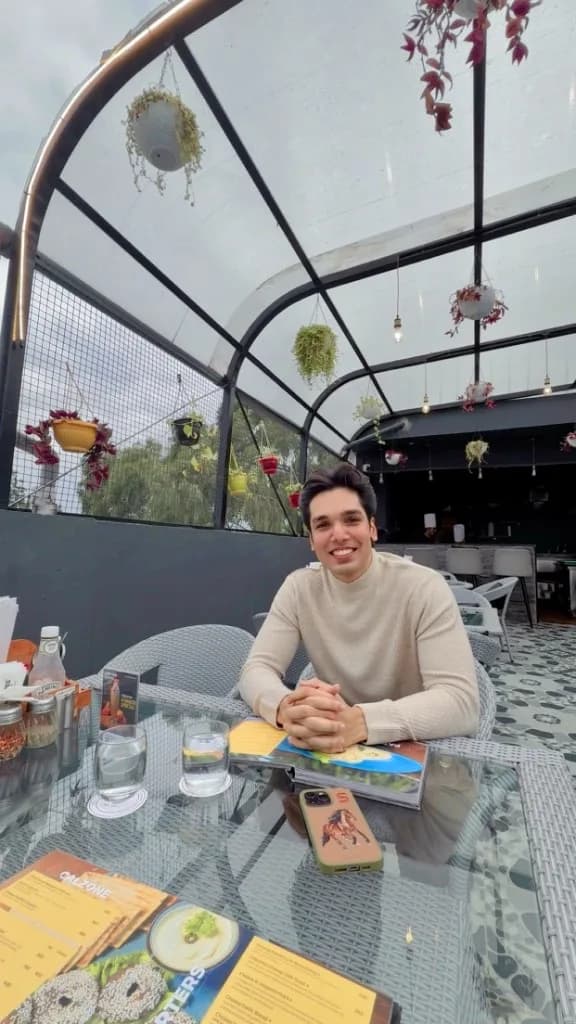 Shivam Tripathi seated at a café table in a cream turtleneck, relaxed posture, looking toward the camera.