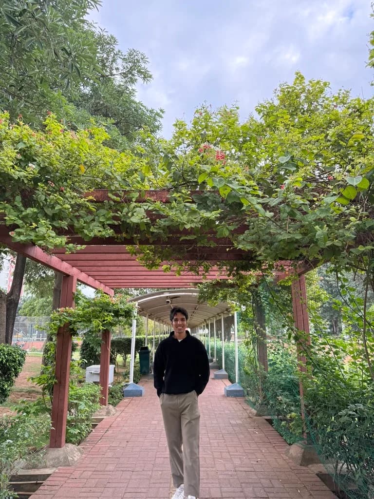 Shivam Tripathi standing outdoors beside a brick path and wooden pergola, smiling at the camera in a casual jacket.